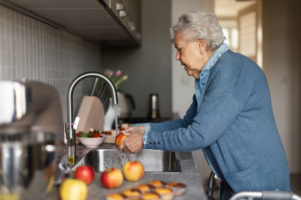 Happy senior woman washing apples in the kitchen. Gradmother is preparing baked goods, muffins, cakes, sweets.