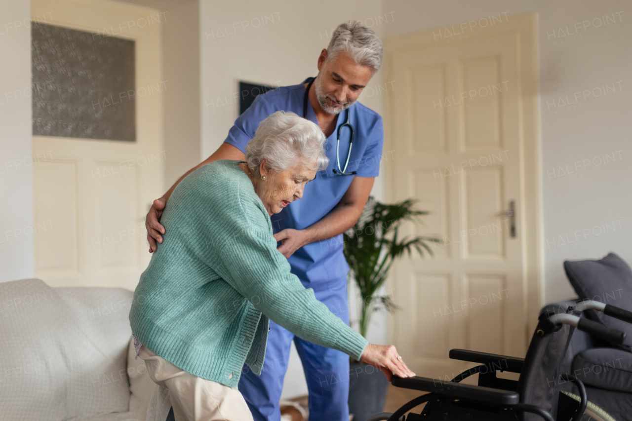 Caregiver helping senior woman to stand from her wheelchair in her home. An elderly woman is patiently trying to get up from her wheelchair.