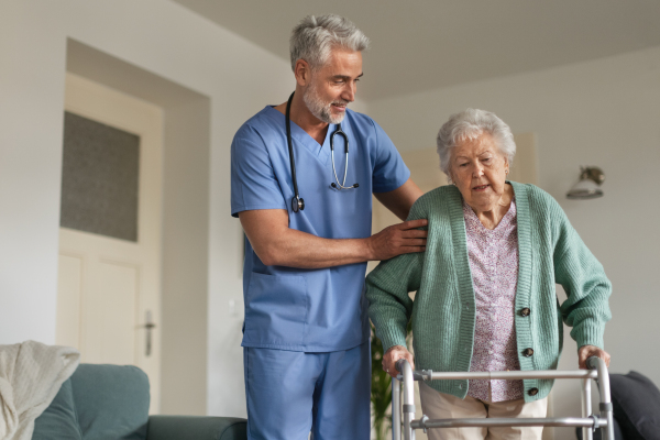 Caregiver helping senior woman to walk in her home. Thoughful male nurse taking care of eldery patient with walker.