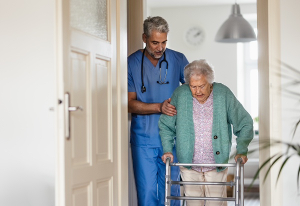 Caregiver helping senior woman to walk in her home. Thoughful male nurse taking care of eldery patient with walker.