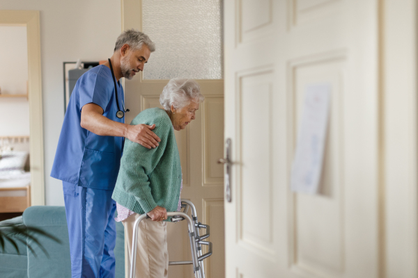 Caregiver helping senior woman to walk in her home. Thoughful male nurse taking care of eldery patient with walker.