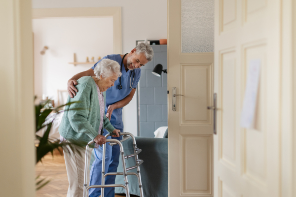 Caregiver helping senior woman to walk in her home. Thoughful male nurse taking care of eldery patient with walker.