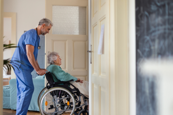 Caregiver pushing senior woman in a wheelchair, helping patient to move in her home. Thoughful male nurse taking care of eldery patient on wheelchair.