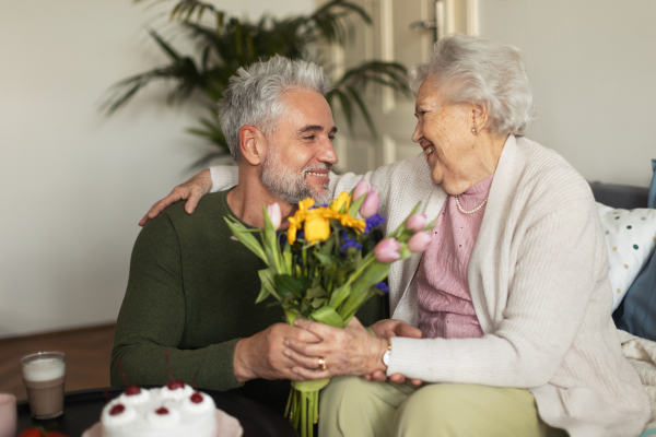 Mature man celebrating with his senior mother. Son giving his mother flowers, gift, cake.