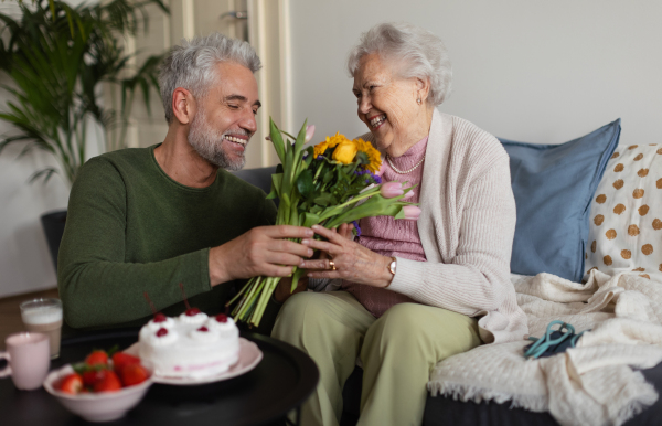 Mature man celebrating with his senior mother. Son giving his mother flowers, gift, cake.