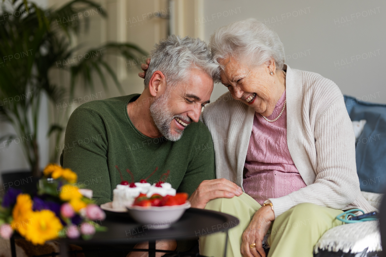 Mature man celebrating with his senior mother. Son giving his mother flowers, gift, cake. Mom and son hugging, touching with foreheads.