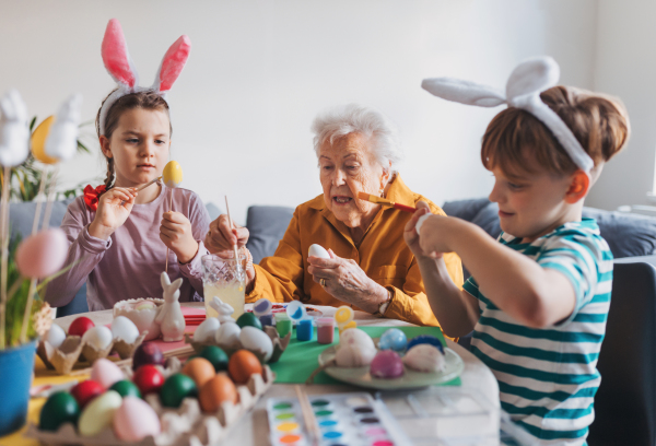 Grandmother with little kids decorating easter eggs at home. Tradition of painting eggs with brush and easter egg dye. Concept of family easter holidays.