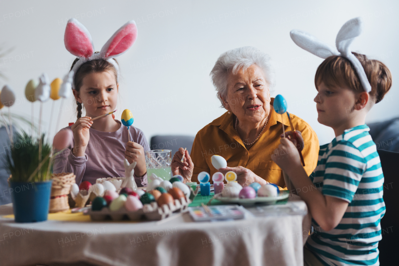 Grandmother with little kids decorating easter eggs at home. Tradition of painting eggs with brush and easter egg dye. Concept of family easter holidays.