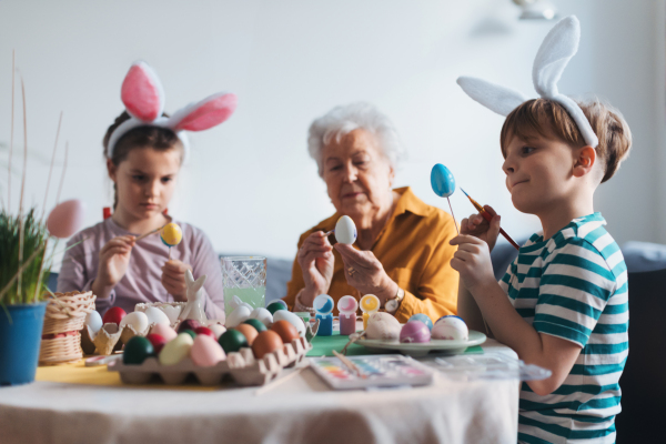 Grandmother with little kids decorating easter eggs at home. Tradition of painting eggs with brush and easter egg dye. Concept of family easter holidays.