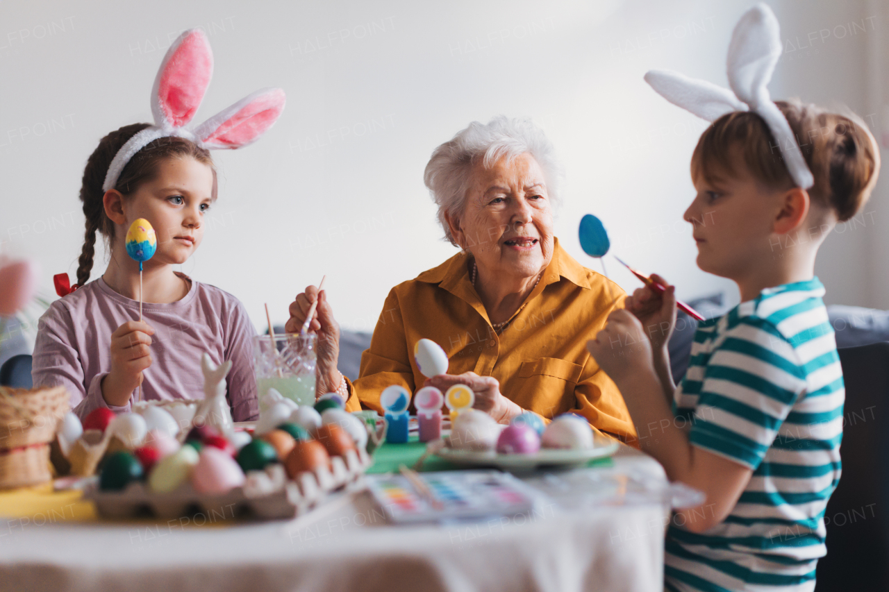 Grandmother with little kids decorating easter eggs at home. Tradition of painting eggs with brush and easter egg dye. Concept of family easter holidays.