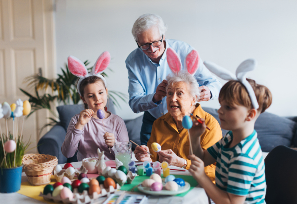 Grandmother with little kids decorating easter eggs at home. Tradition of painting eggs with brush and easter egg dye. Concept of family easter holidays.