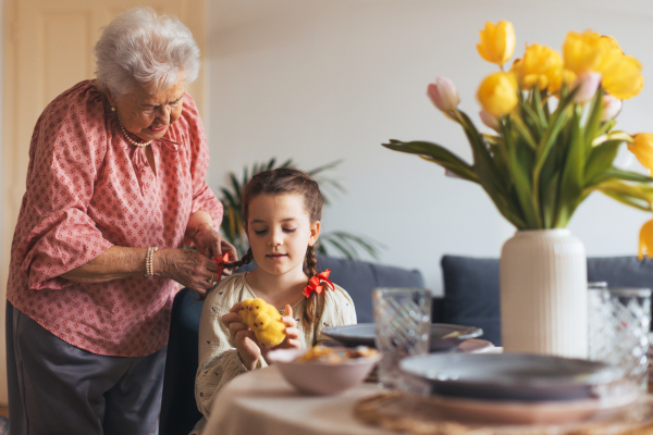 Grandmother braiding hair of granddaughter, putting ribbon ob braid. Prepairing for easter lunch.. Concept of family easter holidays.