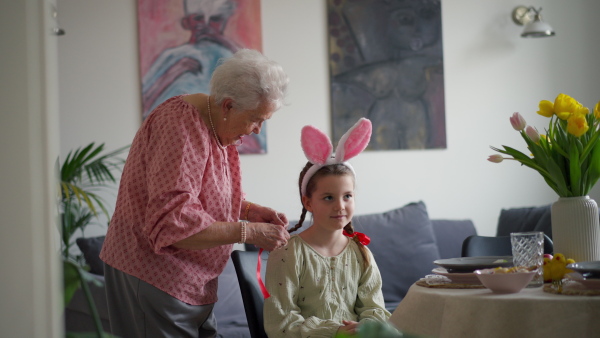 Grandmother braiding hair of granddaughter, putting ribbon ob braid. Prepairing for easter lunch.. Concept of family easter holidays.