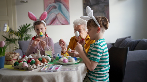 Grandmother with little kids decorating easter eggs at home. Tradition of painting eggs with brush and easter egg dye. Concept of family easter holidays.