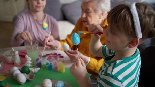 Grandmother with little kids decorating easter eggs at home. Tradition of painting eggs with brush and easter egg dye. Concept of family easter holidays.