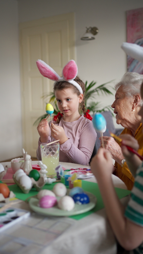 Grandmother with little kids decorating easter eggs at home. Tradition of painting eggs with brush and easter egg dye. Concept of family easter holidays.