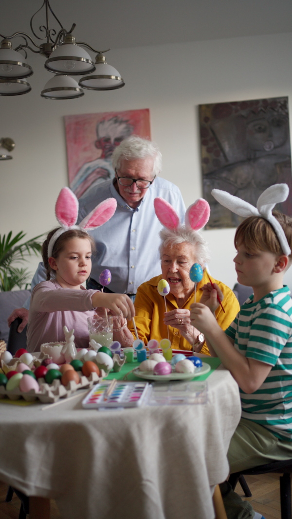 Grandmother with little kids decorating easter eggs at home. Tradition of painting eggs with brush and easter egg dye. Concept of family easter holidays.