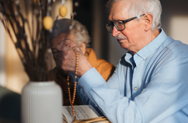 Senior man praying during Easter with the Bible on table, holding rosary, counting prayers, Hail Mary. Preparing for church serice. Easter as religious holiday in Christianity.