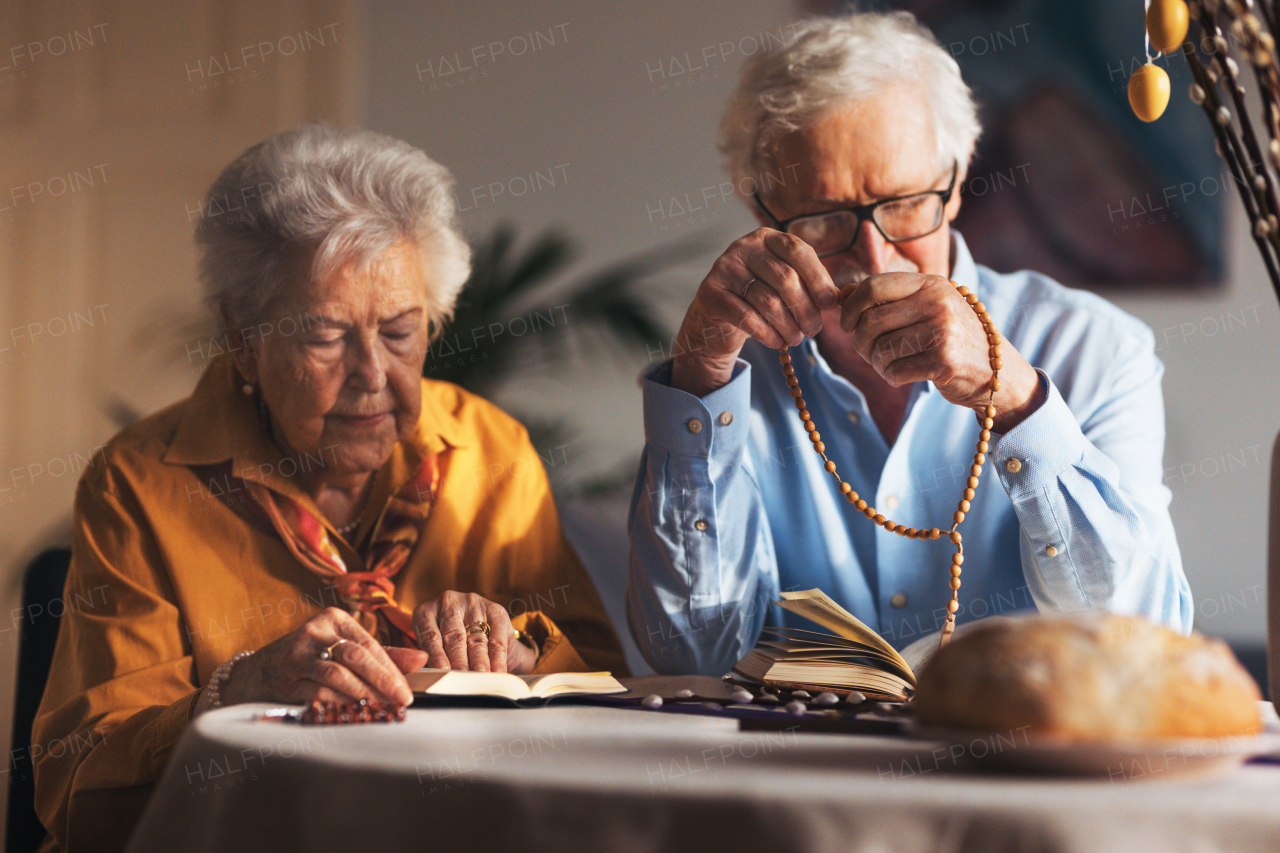 Senior couple praying during Easter, the bible on table at home. Elderly man holding rosary, counting prayers, Hail Mary. Preparing for church serice. Easter as religious holiday in Christianity.