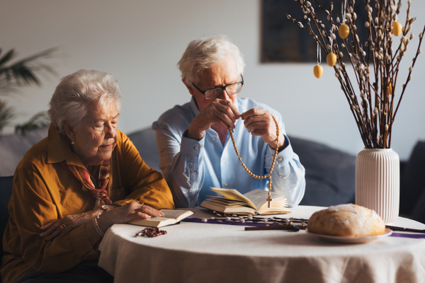 Senior couple praying during Easter, the bible on table at home. Elderly man holding rosary, counting prayers, Hail Mary. Preparing for church serice. Easter as religious holiday in Christianity.