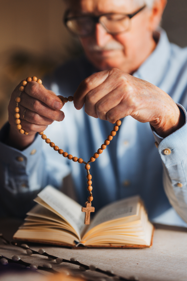 Senior man praying during Easter with the Bible on table, holding rosary, counting prayers, Hail Mary. Preparing for church serice. Easter as religious holiday in Christianity.