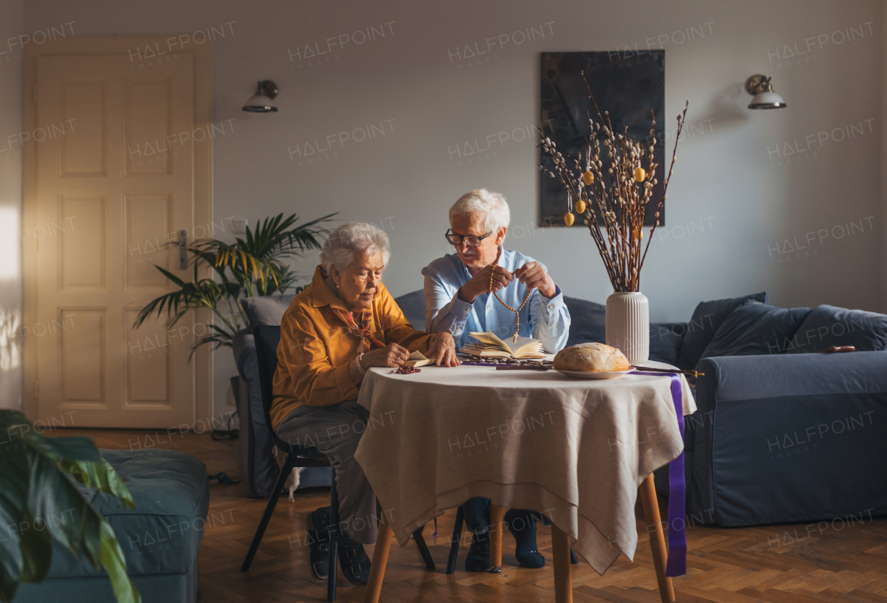 Senior couple praying during Easter, the bible on table at home. Elderly man holding rosary, counting prayers, Hail Mary. Preparing for church serice. Easter as religious holiday in Christianity.