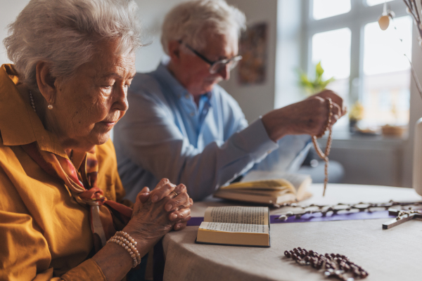 Senior couple praying during Easter, the bible on table at home. Elderly man holding rosary, counting prayers, Hail Mary. Preparing for church serice. Easter as religious holiday in Christianity. Side view.