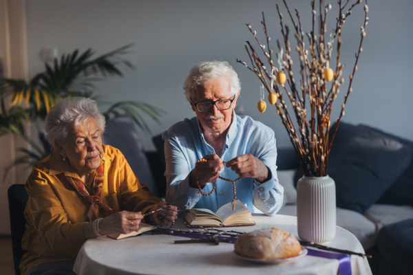 Senior couple praying during Easter, the bible on table at home. Holding rosary, counting Hail Mary prayers. Preparing for church serice. Easter as religious holiday in Christianity.