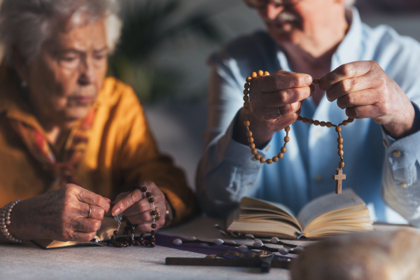 Senior couple praying during Easter, the bible on table at home. Holding rosary, counting Hail Mary prayers. Preparing for church serice. Easter as religious holiday in Christianity. Close up.