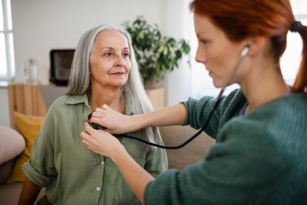 Close-up of nurse examining senior patient with stethoscope at home. Female doctor listening breathing and heart of elderly patient. Check-up after heart surgery.