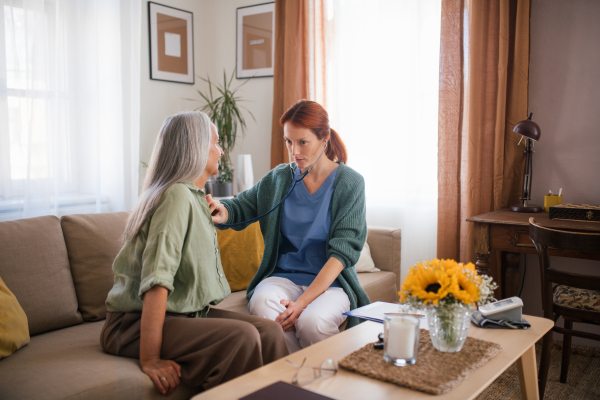 Thoughful nurse examining senior patient with stethoscope at home. Female doctor listening breathing and heart of elderly patient. Check-up after heart surgery.