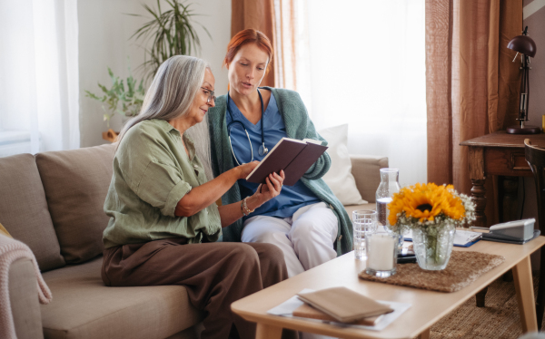 Senior woman and nurse reading book together, having nice time at home.