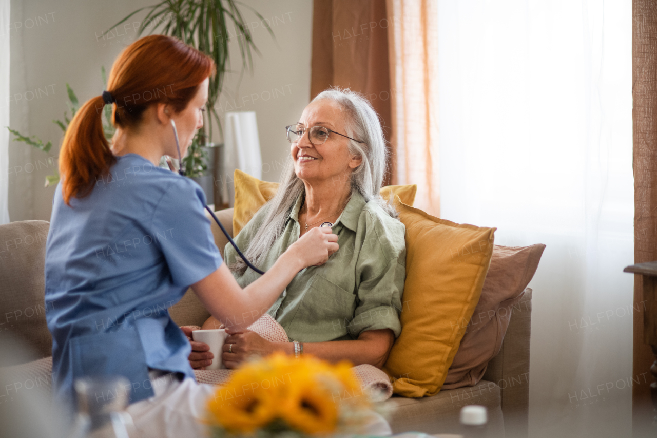 Thoughful nurse examining senior patient with stethoscope at home. Female doctor listening breathing and heart of elderly patient. Check-up after heart surgery.