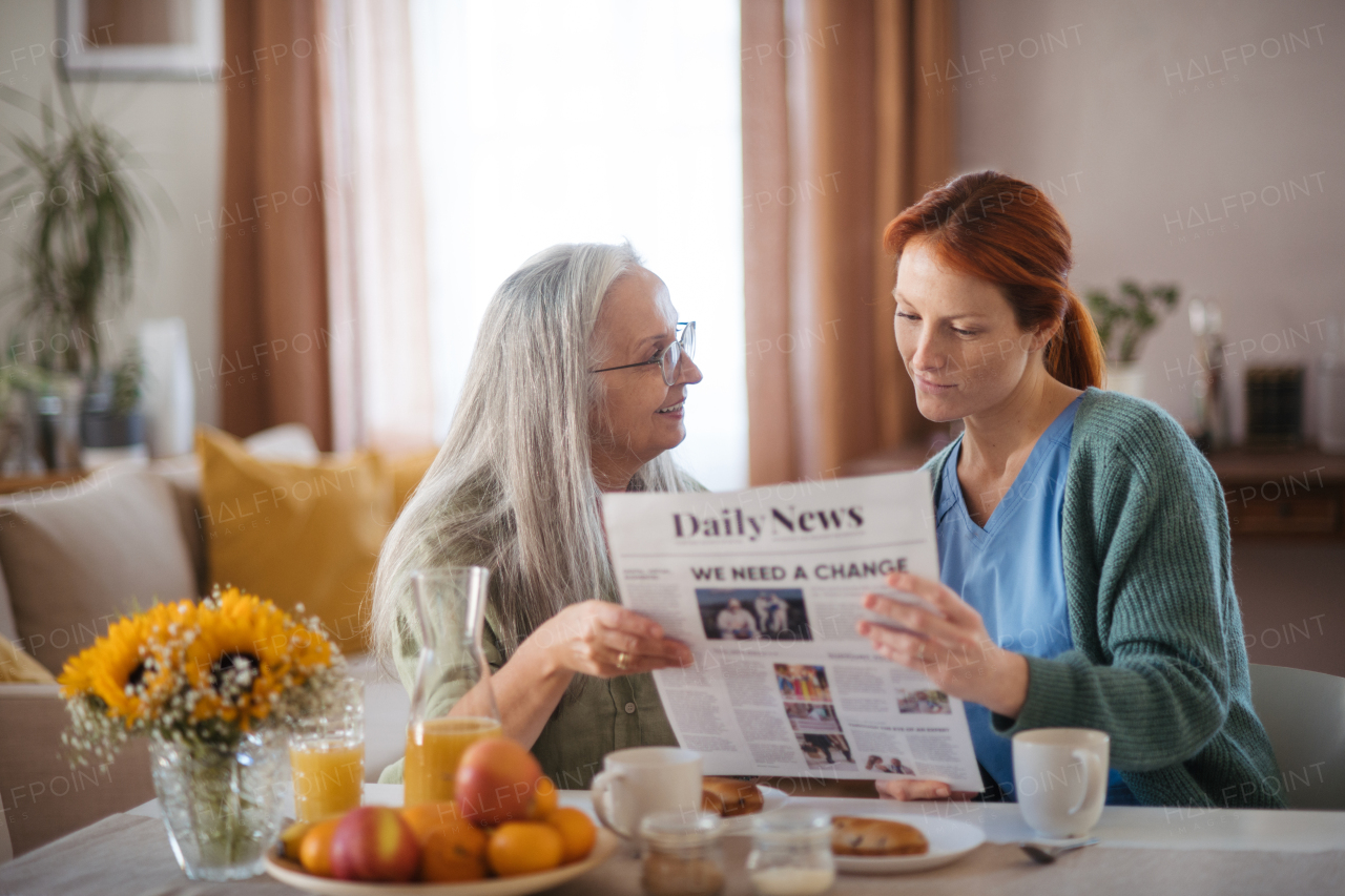 Nurse spending quality time with senior woman at her home, reading newspaper together, discussing latest news. Female caregiver taking care of elderly patient.