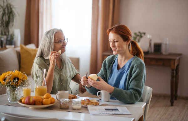 Nurse spending quality time with senior woman at her home, having healthy breakfast together. Female caregiver taking care of elderly patient.