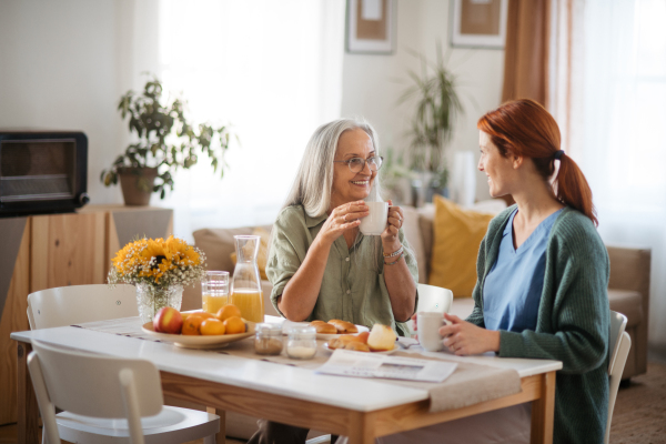 Nurse spending quality time with senior woman at her home, talking and drinking coffee together. Female caregiver taking care of elderly patient.