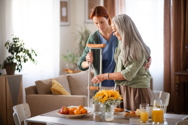 Caregiver helping senior woman to walk with crutches in her home. Thoughful nurse taking care of eldery patient after surgery.