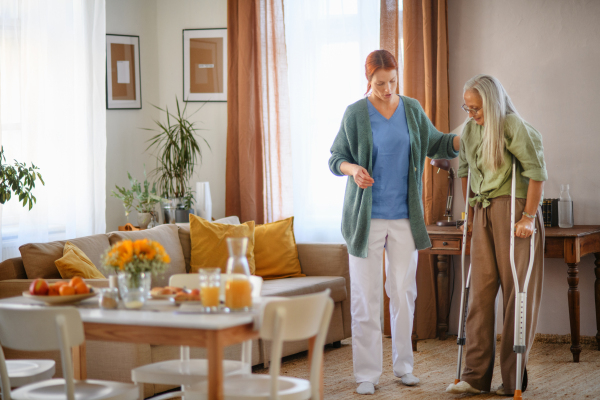 Nurse helping senior woman with walking after leg injury, in her home.