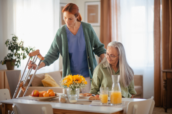 Nurse helping senior woman with walking after leg injury, in her home.