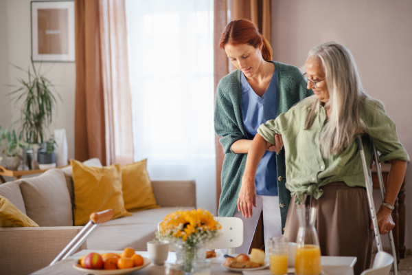 Nurse helping senior woman with walking after leg injury, in her home.