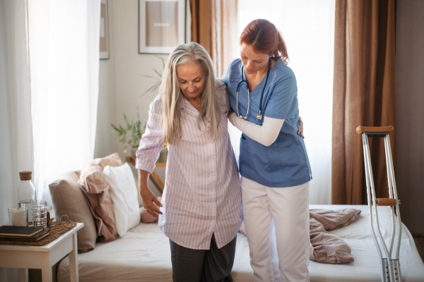 Nurse helping senior woman with walking after leg injury, in her home.