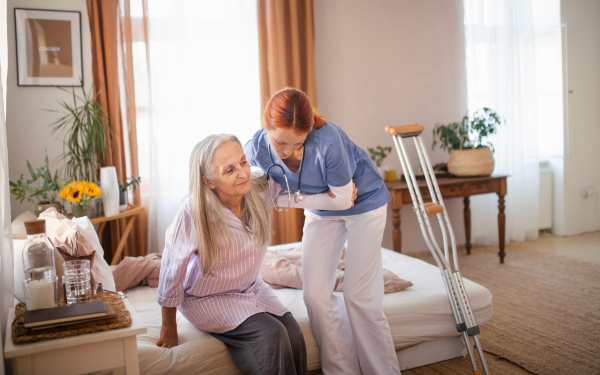 Nurse helping senior woman with walking after leg injury, in her home.