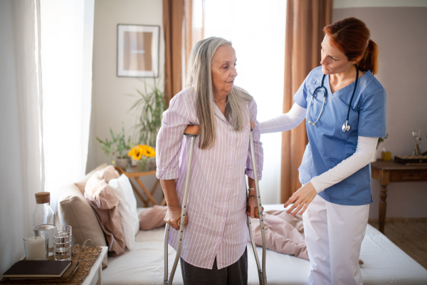 Caregiver helping senior woman to walk with crutches in her home. Thoughful nurse taking care of eldery patient after surgery.