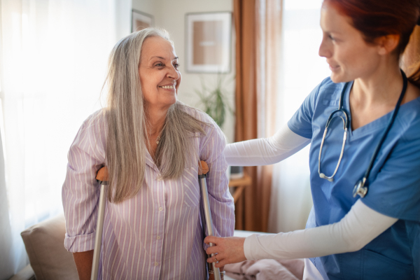 Nurse helping senior woman with walking after leg injury, in her home.