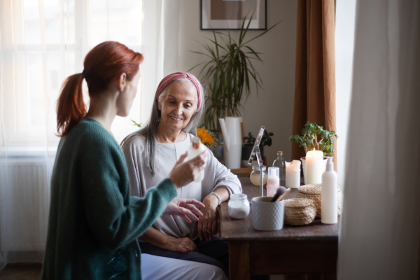 Mature daughter helping her mother with skincare routine. Nurse spending quality time with senior woman at her home, helping her with skincare routine. Woman selling cosmetics product to elderly woman.