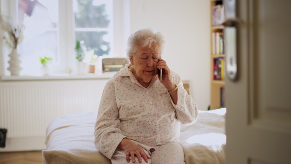 A senior woman crying. Elderly lady has headache, feeling dizzy after waking up. Elderly woman in pyjamas sitting on bed, holding her head.