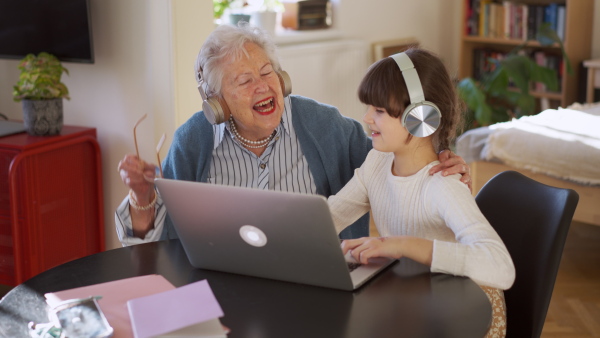 A video of elderly woman spending time with granddaughter. Grandmother with cute girl making video call, wearing headphones, working on laptop.