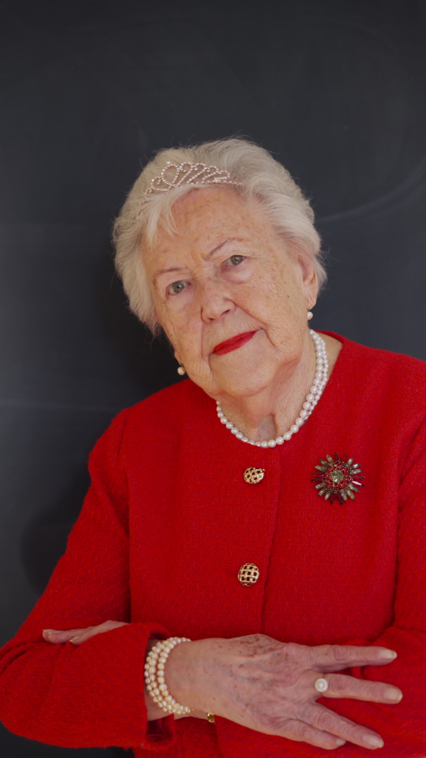 A video of elegant elderly woman with crown in red jacket looking at camera, smiling. Queen lookalike.