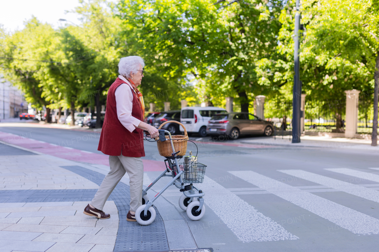 Beautiful senior woman on walk through the city, walking with the help of a rollator. Crossing the crosswalk.