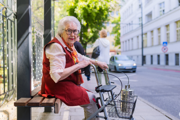 Senior woman in city, sitting on a bus stop, waiting for public transport, rollator next to her.